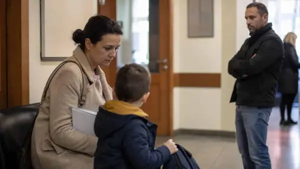 Rodzina w sądzie, matka z synem siedzą, ojciec stoi z boku. Sytuacja sugeruje wniosek o ustalenie kontaktów z dzieckiem.