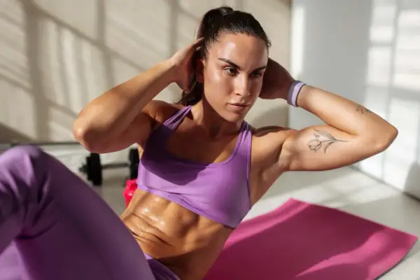 Frau macht Crunches auf einer rosa Matte, um einen flachen Bauch zu bekommen. Sie trägt lila Sportkleidung und hat ein Tattoo auf dem Arm.