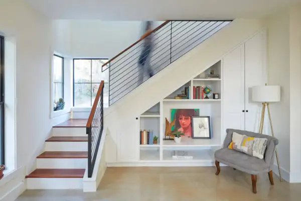 A person ascends a modern staircase, its sleek design contrasting with the built-in bookshelves and storage beneath. This stairwell is a functional art piece.