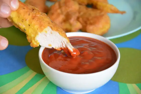 Chrupiący nugget zanurza się w gęstym, czerwonym sosie do nuggetsów. W tle widać talerz z innymi nuggetami.
