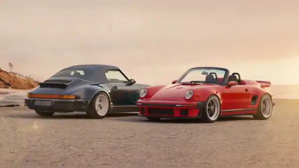 Dos Porsche descapotables, uno rojo y otro azul oscuro, posan en la playa. El **Singer Porsche** rojo destaca con su diseño deportivo y el azul con su capota puesta.