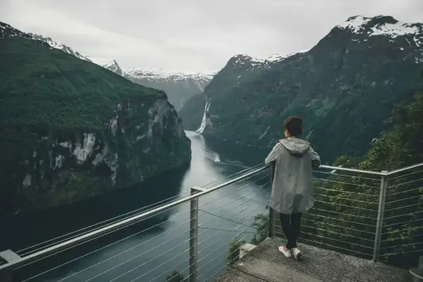 Kobieta podziwia majestatyczne fiordy norweskie. Woda odbija strome, zielone zbocza gór, a w oddali widać wodospad.