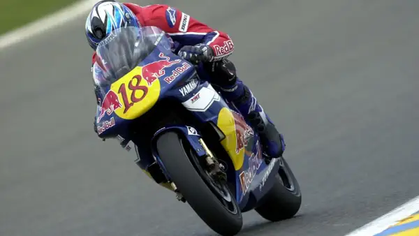 Piloto con casco azul y rojo, traje de carreras Red Bull y moto Yamaha número 18, en plena curva. Jean Michel Bayle en acción.