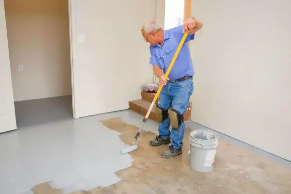 Un homme en bleu peint le sol en béton avec un rouleau. Il porte des genouillères pour peindre le béton confortablement.