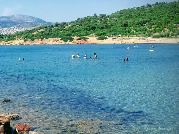 Słoneczna **Ateny plaża** z turkusową wodą, gdzie ludzie kąpią się i relaksują. W tle zielone wzgórza i zabudowania.