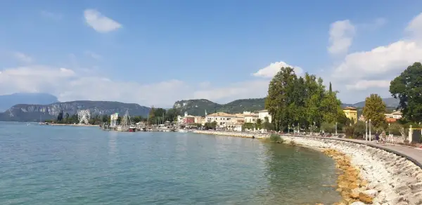 Il lungolago di Bardolino, con il lago azzurro, le barche, le case colorate e le montagne sullo sfondo. Una ruota panoramica si staglia all'orizzonte.