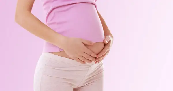 Kobieta w różowej bluzce i beżowych legginsach trzyma dłonie na lekko zaokrąglonym brzuchu. To może być jeden z pierwszych objawów ciąży, jak zmiany w śluzie.