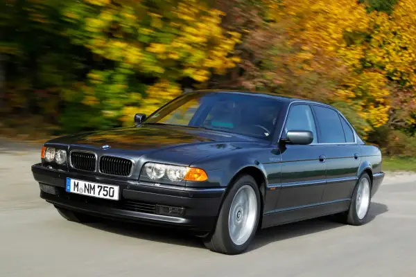 Ciemnoszary BMW E38 Long sunie po drodze, otoczony jesiennymi drzewami w pełnym kolorze.