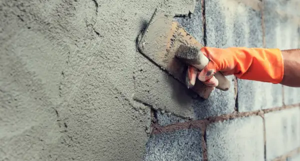 Mano con guanto arancione stende malta da costruzione su un muro di blocchi di cemento con una cazzuola.