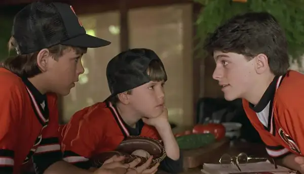 Trzej chłopcy w strojach baseballowych, przypominający scenę z filmów dla dzieci z lat 90. Jeden z nich trzyma rękawicę z piłką.