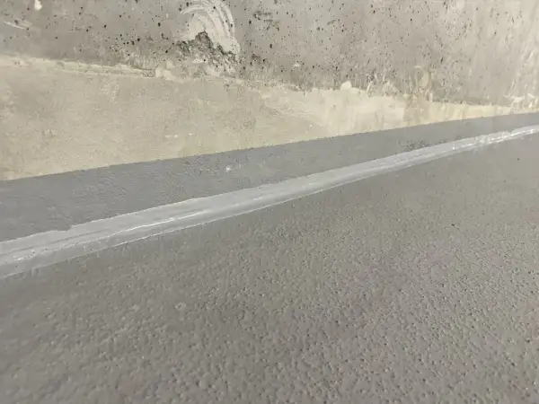 Szara posadzka z widoczną dylatacją wypełnioną białym materiałem, granicząca z betonową ścianą.