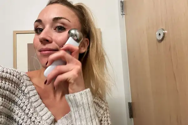 Kvinna med blont hår använder en vit och silverfärgad **ice roller ansikte** för att massera kinden.