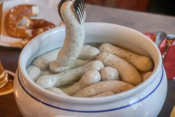 Eine Gabel hebt eine Weißwurst aus einem Topf, um sie zuzubereiten. Daneben liegt eine Brezel.