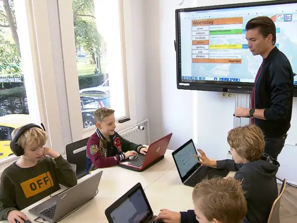 Kinderen leren op laptops in een klaslokaal van een particuliere basisschool. Een leraar staat bij een digibord.