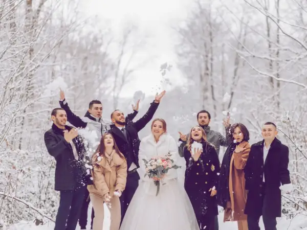 Brautpaar und Gäste werfen Schnee in der Luft. Perfekt für eine Hochzeit im Winter: Was anziehen? Warme Mäntel und festliche Kleidung.