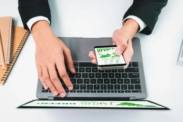 Dłonie na laptopie i smartfonie z wykresami, obok notatniki.