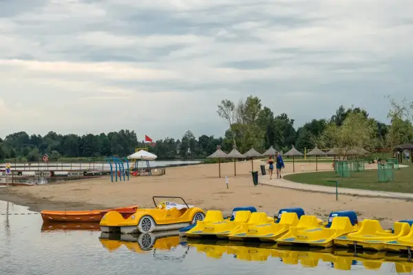 Żółte rowerki wodne i łódki czekają na plaży nad jeziorem blisko Warszawy. Para spaceruje po piasku pod parasolami.