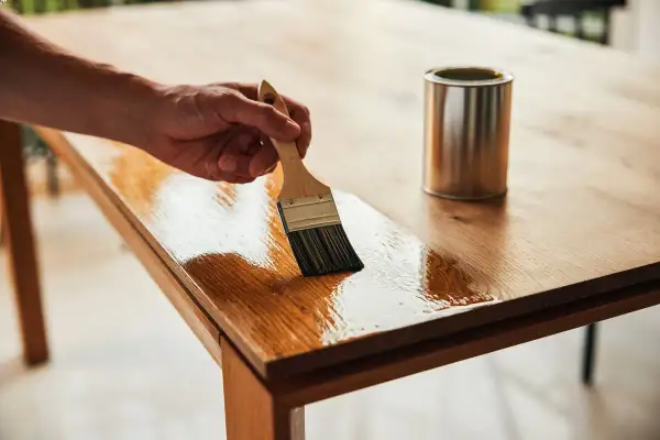 Main d'un artisan appliquant un vernis pour restaurer une table en contreplaqué. La finition brillante redonne vie au bois.