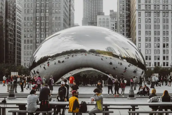 A crowd gathers around Cloud Gate, a reflective public art installation in Chicago, mirroring the city's skyscrapers and sky.