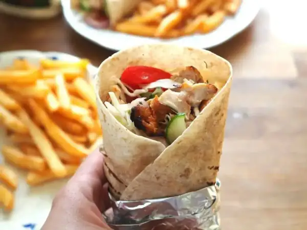 Ręka trzymająca prawdziwy kebab z kurczakiem, warzywami i frytkami w tle.