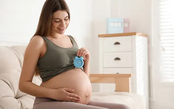 Uśmiechnięta kobieta w 9 miesiącu ciąży trzyma niebieski budzik na brzuchu. Czas na poród!