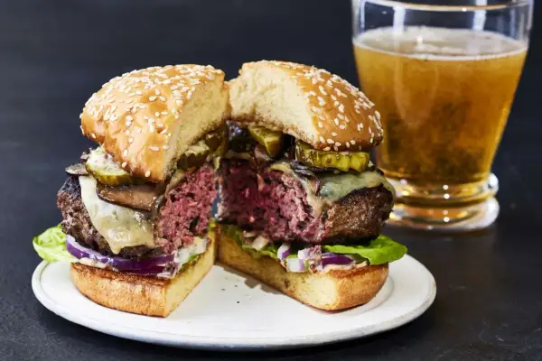 Przekrojony soczysty burger z serem, ogórkiem i grzybami, obok szklanka piwa.