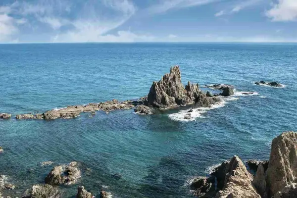 Formaciones rocosas emergen del mar turquesa en la **sierra de Cabo de Gata**. El sol ilumina las aguas cristalinas y el cielo azul con nubes blancas.