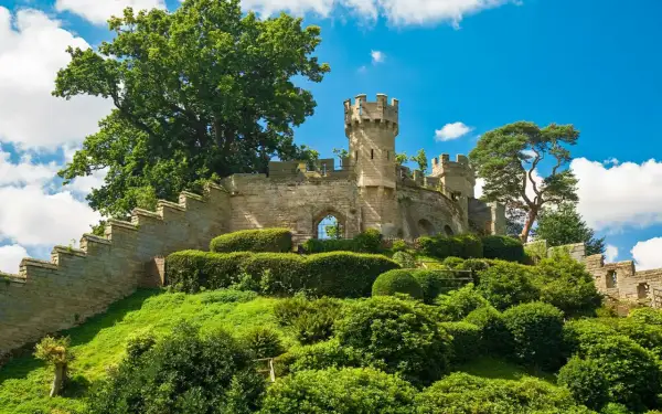 Warwick Castle reser sig stolt på en grönskande kulle, omgiven av frodig grönska och en klarblå himmel med vita moln.