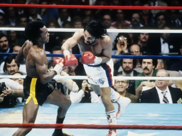 Sugar Ray Leonard lands a punch on Roberto Duran in a fierce boxing match. The crowd watches intently as the two legends battle in the ring.