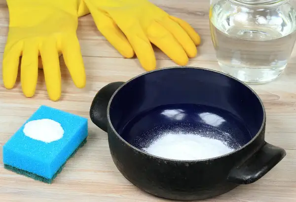 Gants jaunes, éponge bleue avec bicarbonate, casserole noire et un bocal de liquide. Astuces pour savoir comment nettoyer une casserole qui a brûlé.