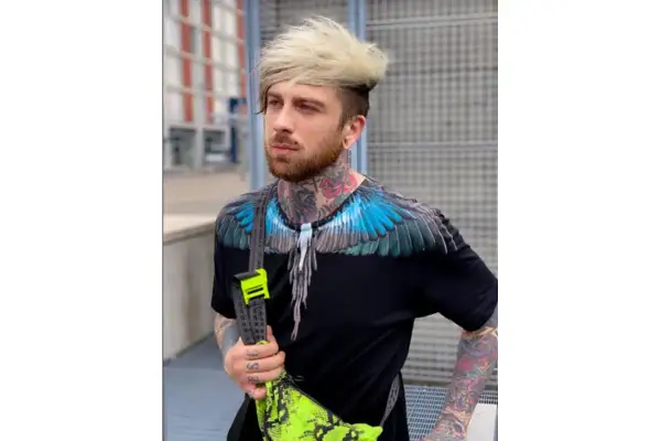 Hombre con tatuajes visibles en cuello y brazos, viste camiseta negra con alas azules y bolso neón.