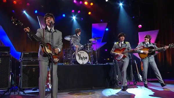 Una cover band sul palco, con abiti grigi e chitarre, ricorda i Fab Four.