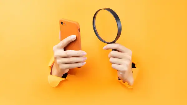 Ręce trzymające pomarańczowy iPhone i lupę, sugerujące, jak namierzyć iPhone po IMEI.