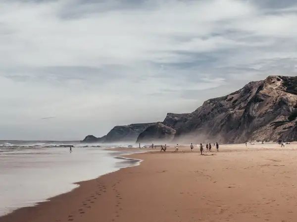 Plaża w Faro, wrzesień. Ludzie grają w piłkę, spacerują po piasku. W tle skaliste klify i ocean.