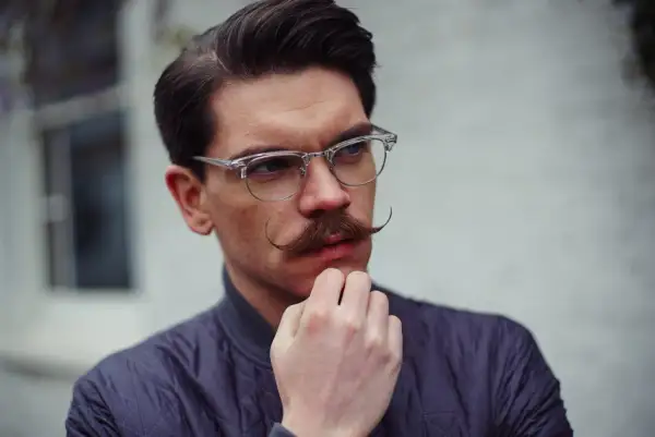 Un homme au visage losange, lunettes fines et moustache élégante, réfléchit.