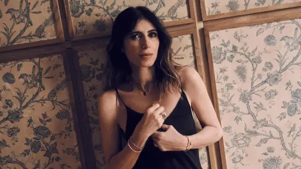 Woman in black slip dress leans against a wall with floral wallpaper.