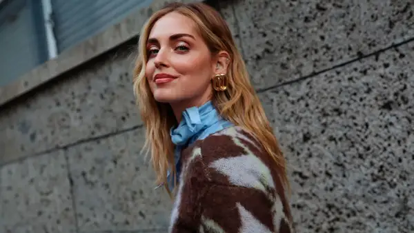 Mujer con media melena rubia, pendientes dorados y jersey estampado, sonriendo ante un muro de hormigón.