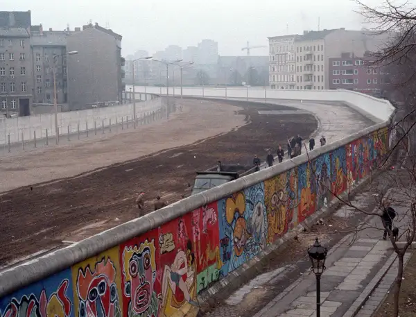 Kolorowy Mur Berliński, świadectwo czasów, kiedy powstał mur berliński, z ludźmi spacerującymi po jego szczycie i budynkami w tle.