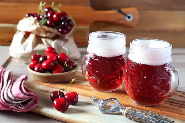 Due boccali di birra kriek rossastra con schiuma bianca, circondati da ciliegie fresche su un tagliere di legno.