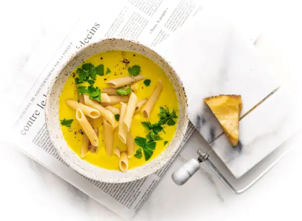 Zupa neapolitańska z makaronem penne i natką pietruszki, podana na gazecie. Obok kawałek sera i serownik.