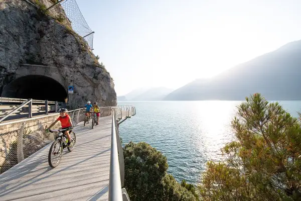 Ciclisti su una pista ciclabile panoramica lungo il lungolago di Desenzano, con un tunnel scavato nella roccia e il lago scintillante.