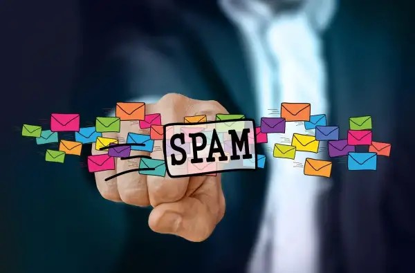 Dłoń wskazuje na napis SPAM otoczony kolorowymi kopertami, symbolizującymi niechciane wiadomości. To definicja spamu.