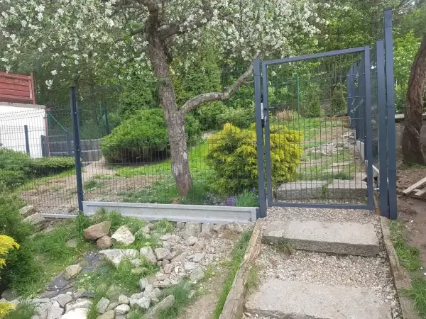 Nowe ogrodzenie z furtką, prawdopodobnie 120 cm wysokości, otacza zielony ogród z kwitnącym drzewem.
