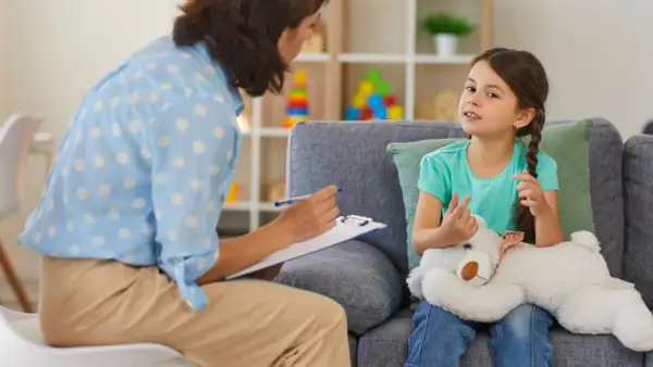 Psychoterapeutka z notatkami rozmawia z dziewczynką trzymającą misia.