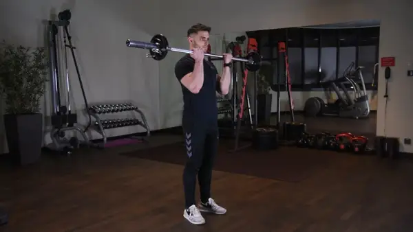 Mężczyzna wykonuje ćwiczenie barbell curl w siłowni. W tle widać sprzęt do ćwiczeń.