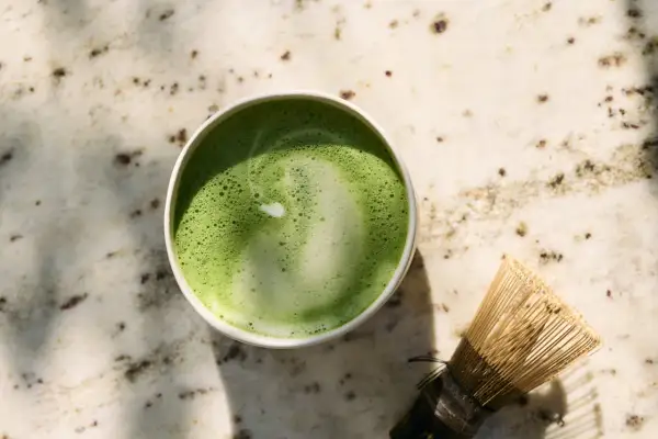 Zielone matcha w filiżance obok bambusowej miotełki na marmurowym blacie.