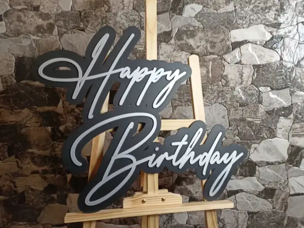 Czarny napis "Happy Birthday" z białymi obrysami, stojący na sztaludze. Idealny na życzenia "sto lat"!