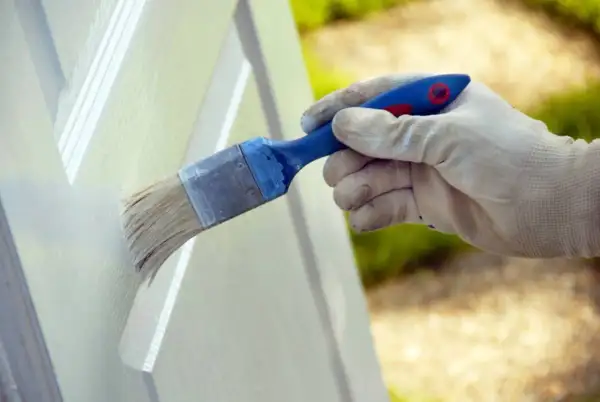 Mano enguantada aplicando pintura blanca a una puerta con brocha. Proceso de lacar puertas en blanco para renovar su aspecto.