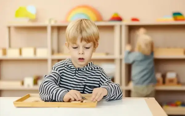 Chłopiec skupiony na zadaniu z materiałami Montessori. W tle inne dziecko i regały z zabawkami.