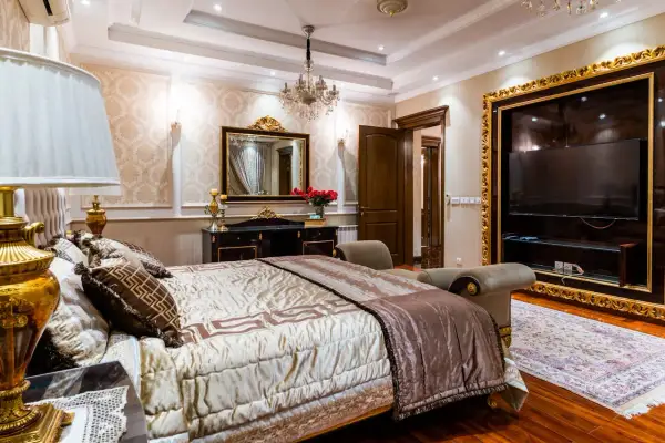 Elegancka sypialnia glamour beżowa z bogato zdobionym łóżkiem, kryształowym żyrandolem i złotymi detalami.